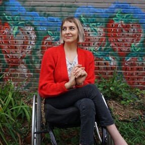 Rebekah Taussig white woman with chin length like colored hair with bangs wearing white top, red sweater and dark pants sitting in wheelchair with legs crossed in front of painted mural of strawberries.acket smiling at camera