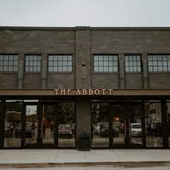 photo of the front entrance of The Abbott event center. Two story gray brown brick with lot of windows and sidewalk in front.